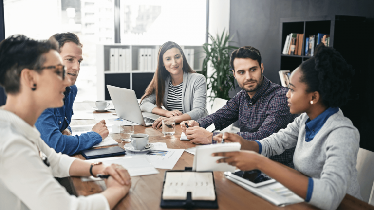 A group of five diverse colleagues in a meeting room engage in discussion. Papers, a laptop, and cups are on the table, conveying collaboration.