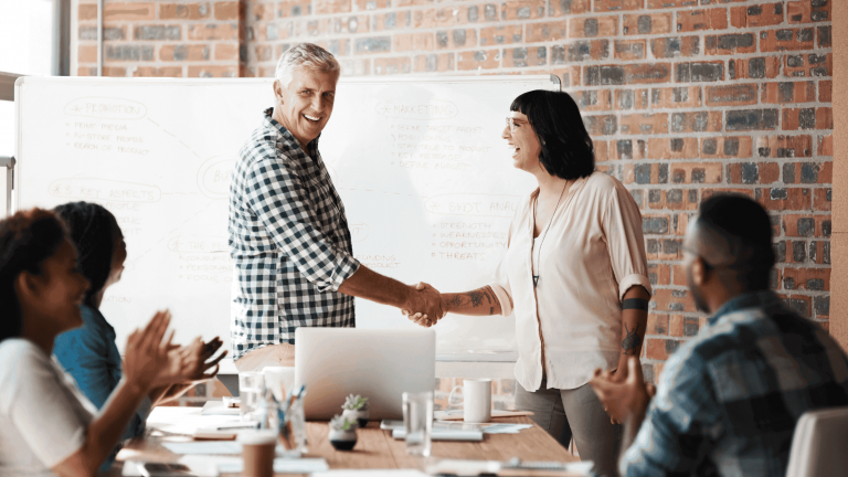 A diverse group in a meeting room, two people smiling and shaking hands, others clapping. A whiteboard with notes is in the background, creating a positive tone.