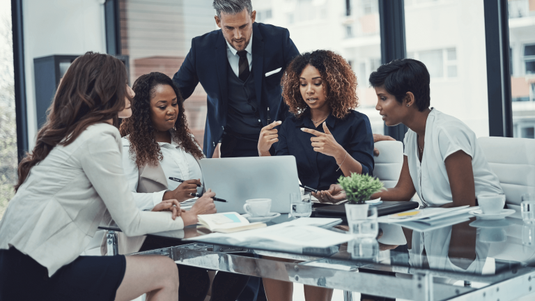 A diverse group of five people in a modern office, actively discussing around a laptop. Papers and coffee cups are on the glass table, conveying teamwork.