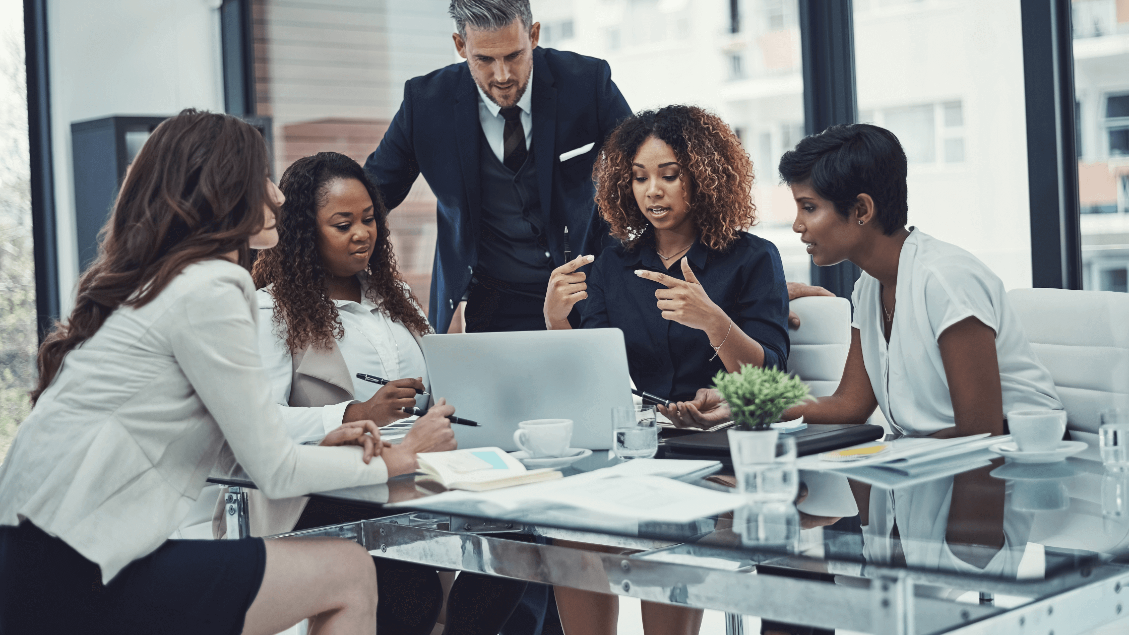 A diverse group of five people in a modern office, actively discussing around a laptop. Papers and coffee cups are on the glass table, conveying teamwork.