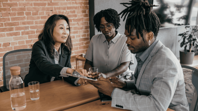 Three people in a business meeting sit at a wooden table with notepads. A woman shows a tablet to a man, while another woman observes, conveying focus.