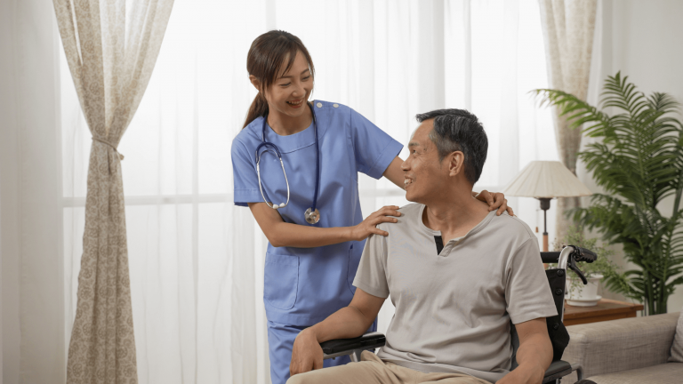 Female nurse in blue scrubs smiles at a man in a wheelchair, placing a hand on his shoulder in a cozy room with light curtains and a plant.