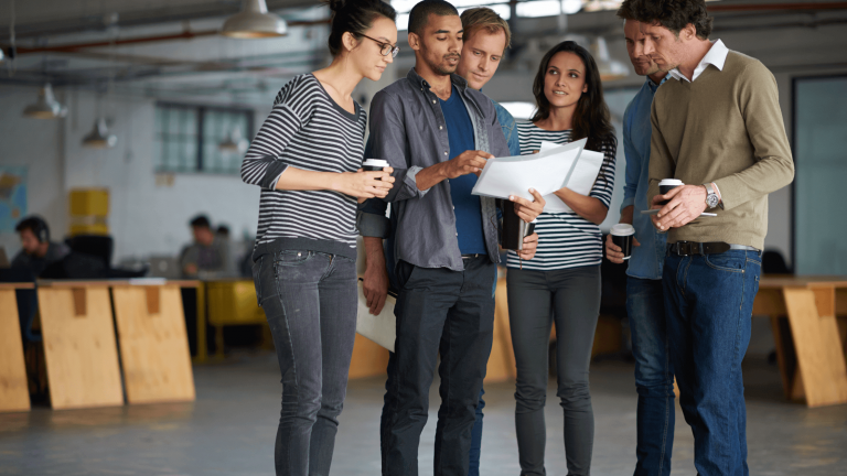 A diverse group of five people in casual attire stand closely, discussing documents in a modern office. They look focused and collaborative.
