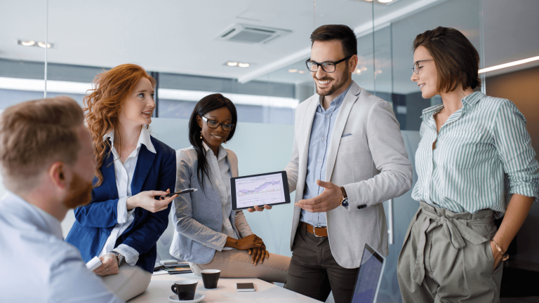 A diverse group of professionals smiling and collaborating in an office. A man holds a tablet showing graphs, suggesting a positive business meeting.