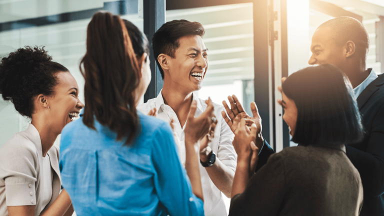 A group of diverse colleagues in an office, smiling and applauding together. The setting is bright and conveys a sense of celebration and teamwork.