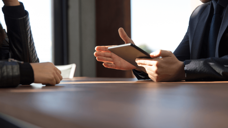 Two people are seated at a wooden table. One holds a tablet, gesturing, while the other listens attentively. The scene suggests a business meeting.