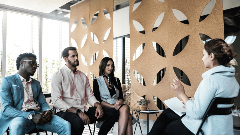 Four people sit in a modern office, engaging in a discussion. The setting is relaxed, with decorative panels and soft natural light streaming in through large windows.