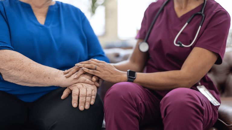 A healthcare worker in burgundy scrubs compassionately holds an elderly person's hand, who is wearing blue. The scene conveys warmth and support.
