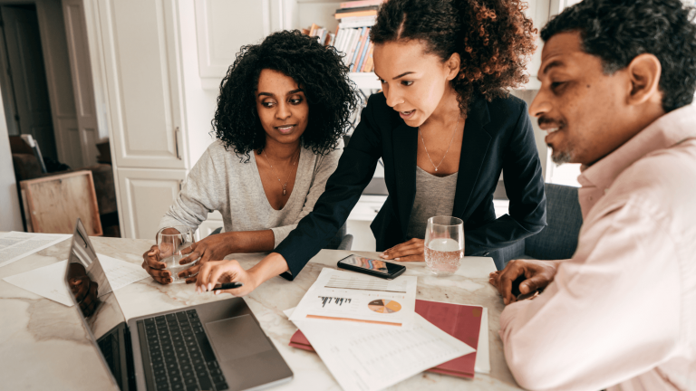 Three people are discussing over a laptop at a table, with documents and a pie chart. The scene conveys focused collaboration and teamwork.