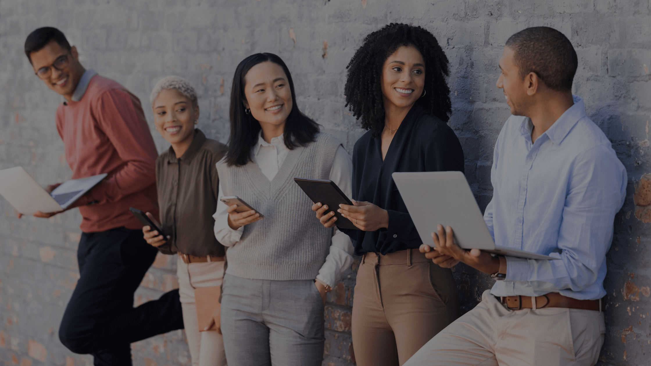 Five diverse individuals stand against a brick wall, smiling and engaging while holding laptops and phones, conveying teamwork and collaboration.