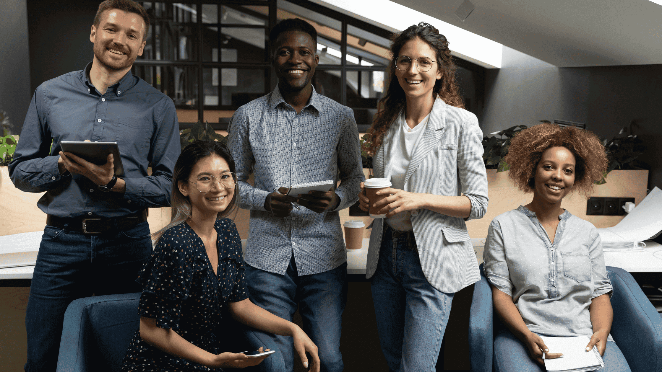 A diverse group of five professionals stands together in an office setting, holding notebooks, tablets, and coffee cups.