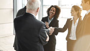 A group of business professionals in suits are smiling, shaking hands, and interacting warmly in a bright office setting, conveying friendliness and success.