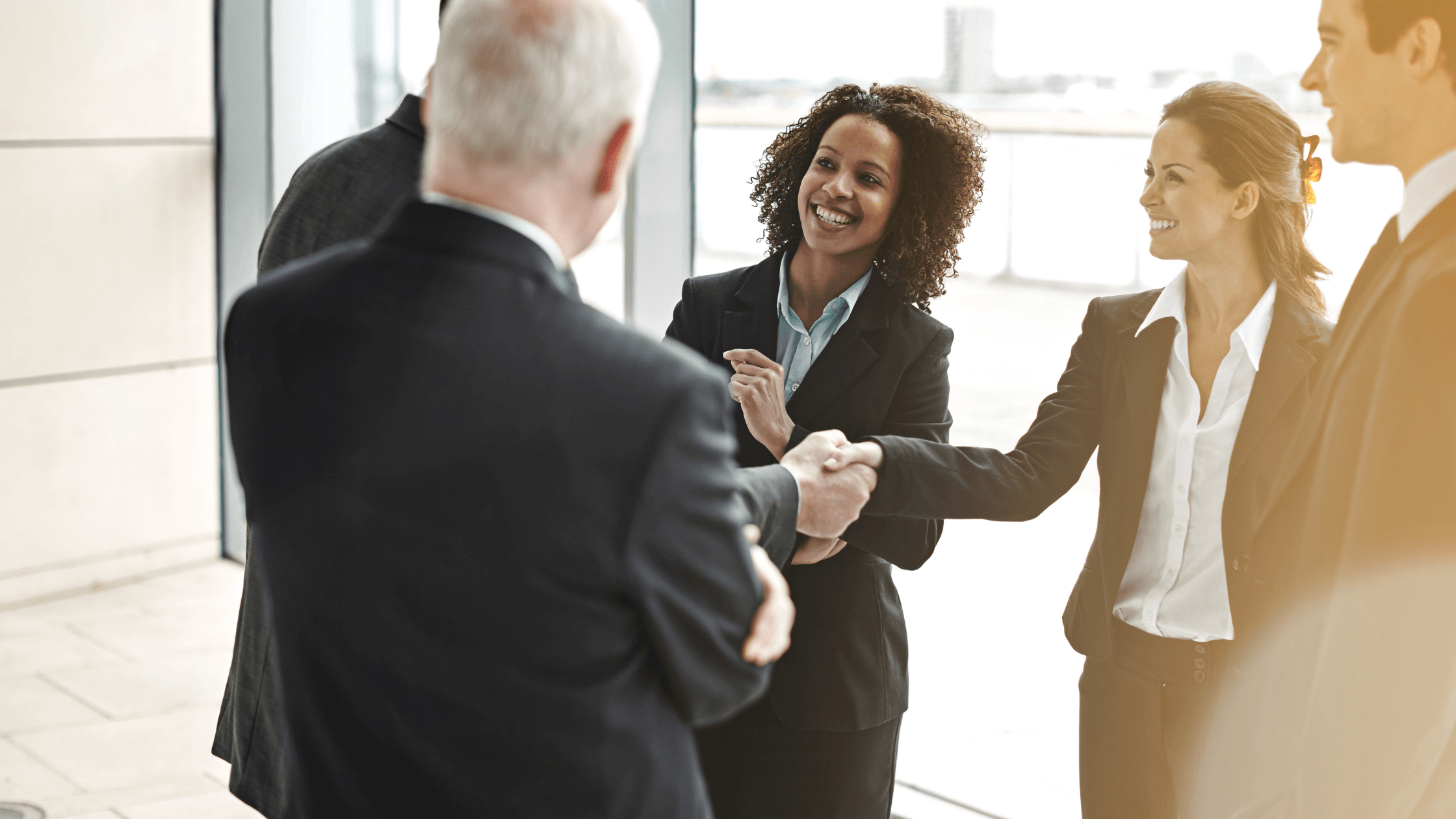 A group of business professionals in suits are smiling, shaking hands, and interacting warmly in a bright office setting, conveying friendliness and success.