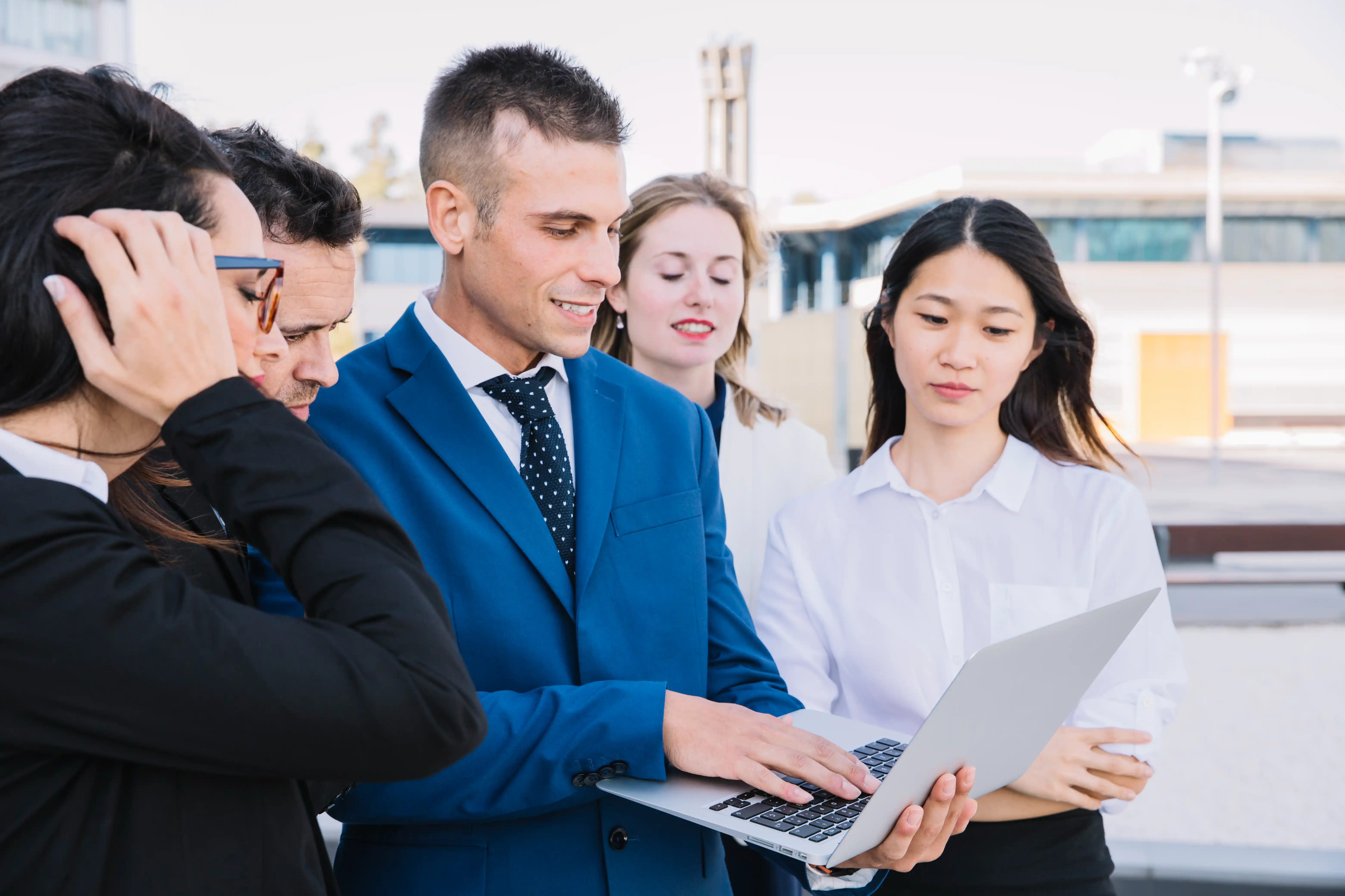 A diverse group of business professionals gathered around a laptop, engaged in discussion and collaboration.