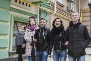 A group of five people walking outdoors, smiling and looking up. They're dressed in casual jackets and scarves, with colorful buildings in the background.