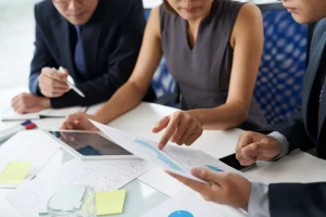 Three people in business attire discuss documents at a table with charts and a tablet, conveying focus and collaboration in a professional setting.