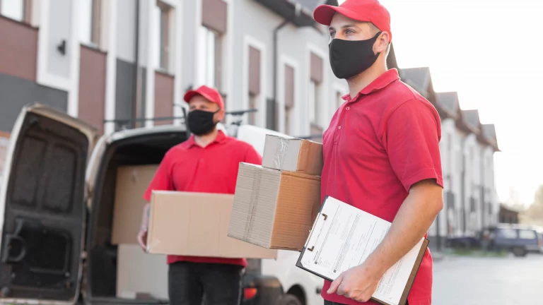 Two delivery workers in red shirts handle packages outside a van, with residential buildings in the background.