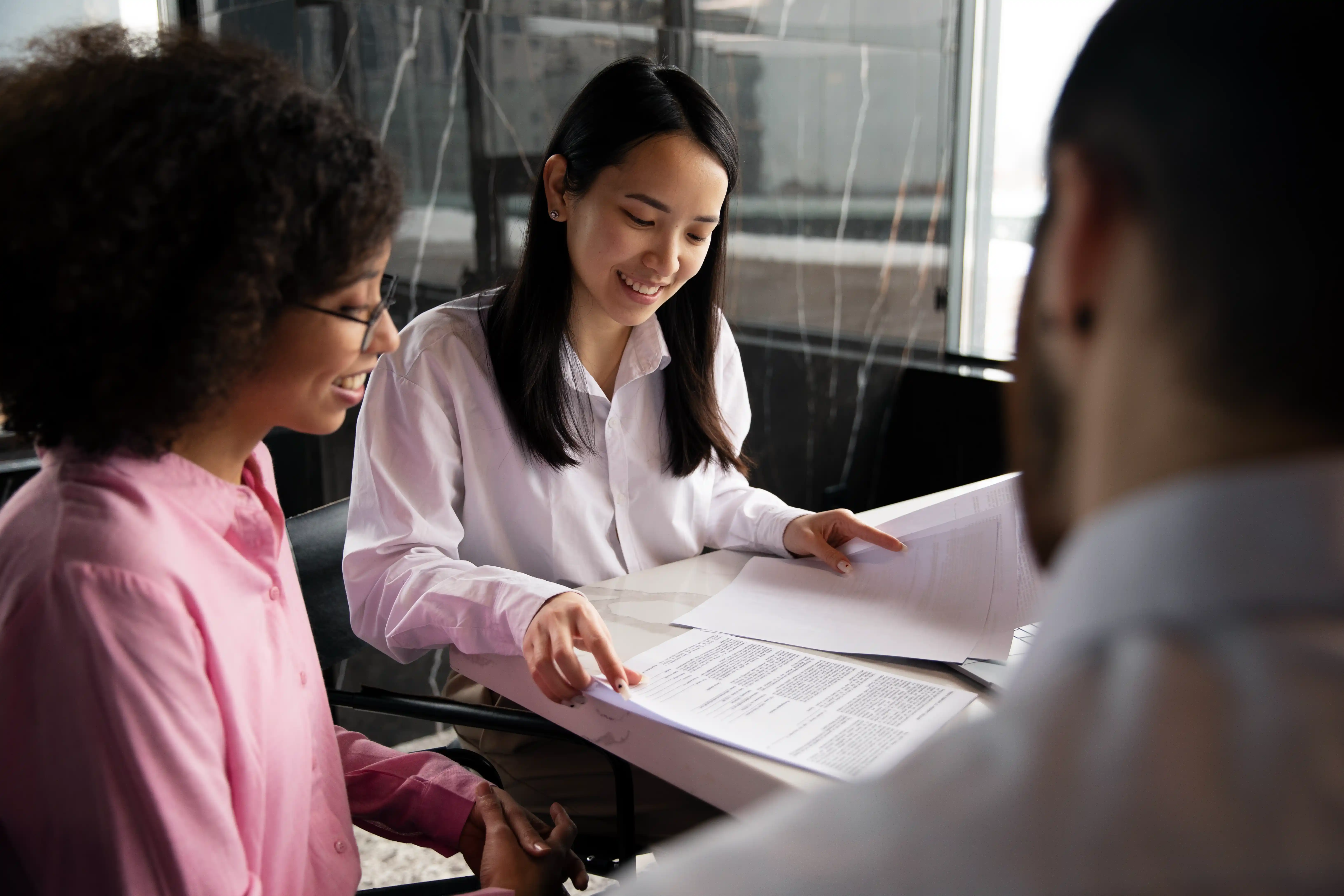 Three people in an office setting discuss documents. A woman smiles and points at a page, conveying a collaborative and positive atmosphere.
