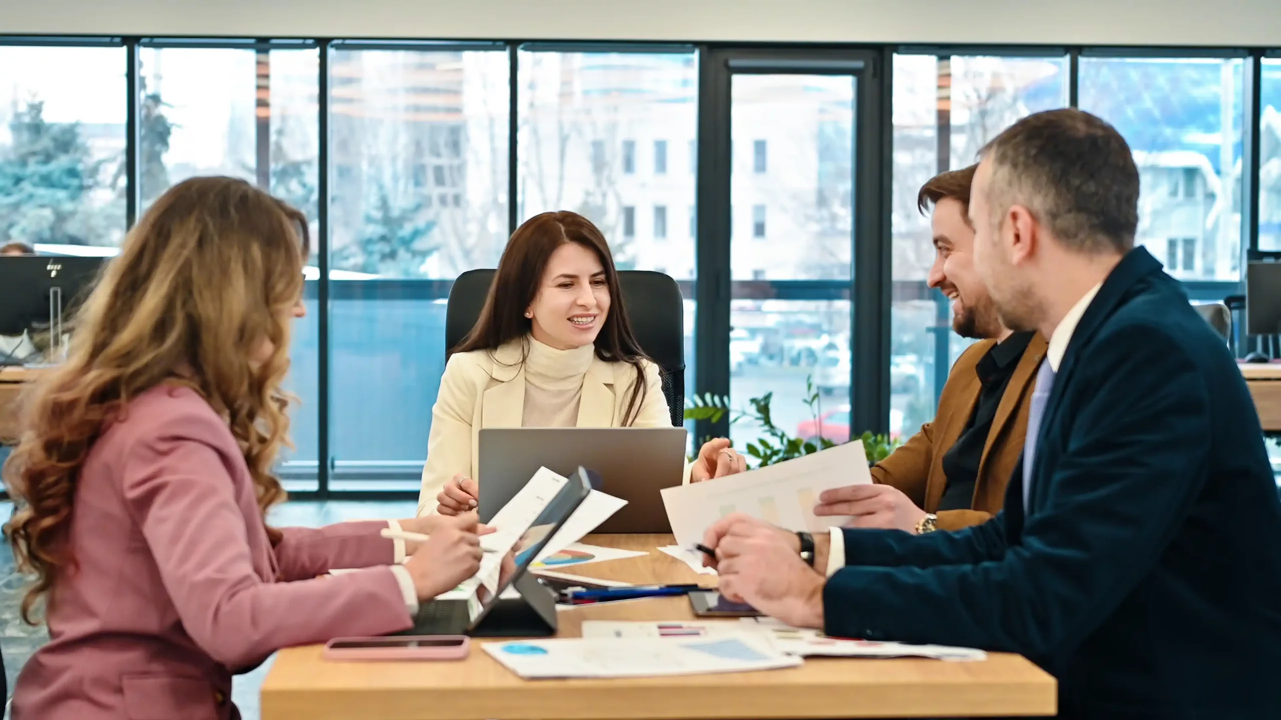 Four people sit at a conference table in a modern office, engaged in discussion. They appear collaborative and focused, with papers and a laptop visible.